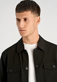 Zwarte jas met knopen en een kraag, voorzien van twee zakken en een gladde textuur, gedragen over een wit overhemd. Simpel, modern ontwerp.