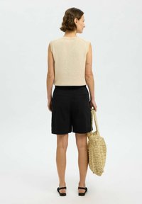 Svetlo bež brezrokavnik z teksturiranim pletenjem, v kombinaciji s črnimi kratkimi hlačami in tkano slamnato torbo. Sandali zaokrožajo opravo.