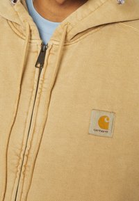 Sudadera con cremallera en color beige, hecha de un tejido texturizado, que presenta un parche de logo visible en el lado izquierdo del pecho y cordones ajustables en la capucha.