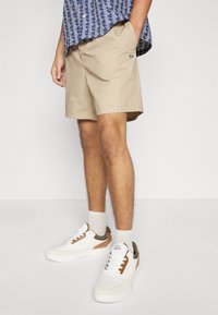 Beiga shorts i lätt tyg, med sidofickor och en liten logotyp. Matchas med vita sneakers och ljusgrå strumpor.