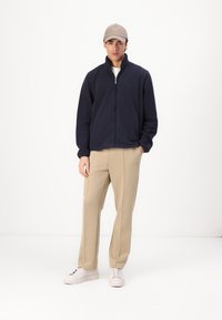 Jachetă din fleece cu fermoar de culoare bleumarin, cu un guler înalt, combinată cu pantaloni bej și adidași albi. Modelul poartă o șapcă de un bej deschis.