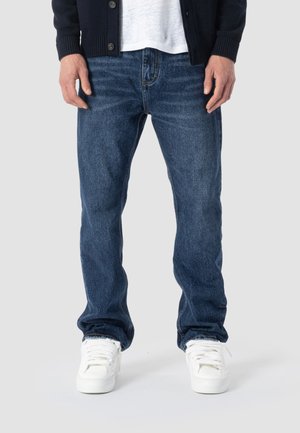 Mand iført mørkeblå jeans med lige ben, hvide sneakers, hvid t-shirt og sort cardigan stående mod en ensfarvet baggrund.