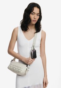 Valge nahkne ristiõlakott, millel on kootud detail. Omab hõbedast metallist aksessuaare ja eemaldatavat metallist kaarditaskut, mis on külge kinnitatud.