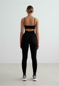 Zwart sportbeha met dunne bandjes, gecombineerd met hoge zwarte leggings. Het model draagt witte sportschoenen met zwarte accenten.