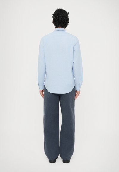 Vaaleansininen pitkähihainen paita, jossa on napillinen kaulus, yhdistettynä väljälahkeisiin, tummanharmaisiin teksturoiduihin housuihin, takaa katsottuna.