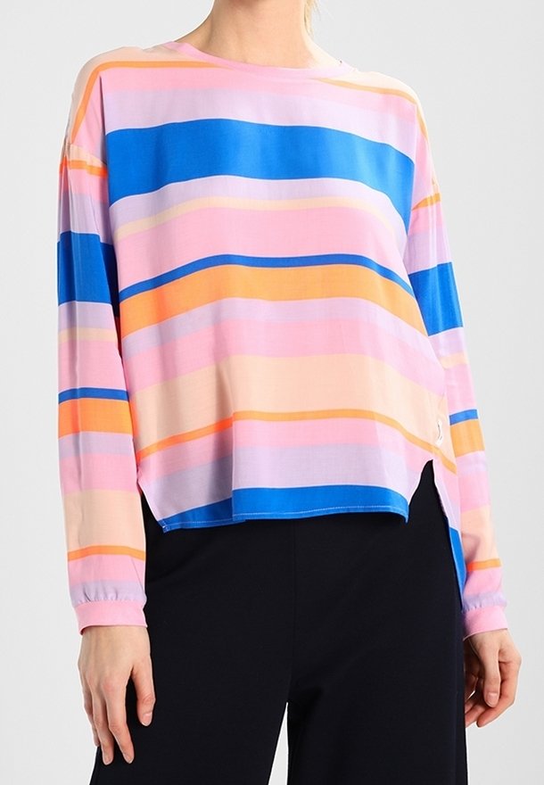Femme portant un haut à manches longues avec des rayures horizontales en rose, bleu, orange et beige, associé à un pantalon noir.