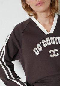 Primer plano de una persona que lleva un sudadera marrón oscuro con rayas blancas en las mangas y el texto blanco "CO' COUT" y el número "33" en el pecho.