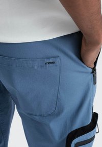 Pantalons bleus avec une poche arrière portant un logo noir et des poches latérales zippées avec des accents noirs, fabriqués en tissu lisse.