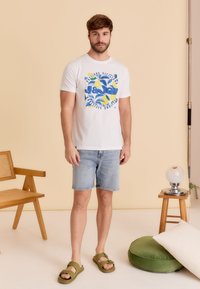 Witte graphic t-shirt met blauwe en gele citruspatronen, gecombineerd met lichte denim shorts en olijfgroene sandalen. Eenvoudig ontwerp, casual stijl.