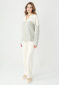 Veste en pull gris clair et crème avec un motif diagonal, fermeture éclair intégrale, col et tissu texturé ; associée à un pantalon crème et des baskets beiges.