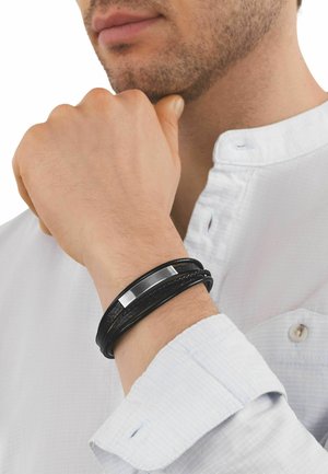 Uomo con camicia bianca che indossa al polso un bracciale in pelle nero a più fili con una piastra centrale in metallo argentato.