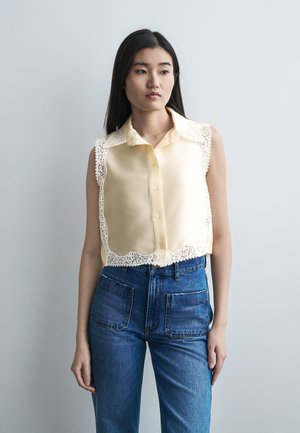 Νεαρή γυναίκα με μακριά σκούρα μαλλιά φοράει αμάνικη ανοιχτό κίτρινη μπλούζα με δαντελωτά άκρα και ψηλόμεσα μπλε τζιν, όρθια μπροστά σε απλό φόντο.