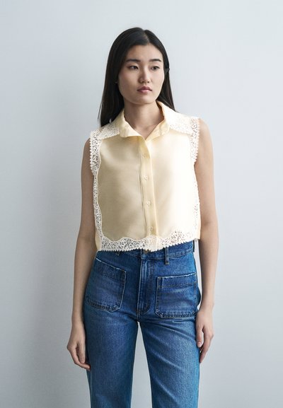 Jeune femme aux longs cheveux noirs portant un chemisier sans manches jaune pâle avec des bords en dentelle et un jean bleu taille haute, se tenant devant un fond uni.