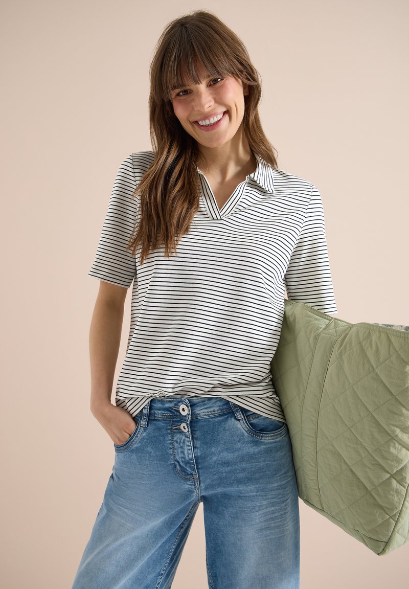 Femme souriante aux longs cheveux bruns portant un polo rayé et un jean bleu, tenant un grand sac cabas matelassé vert sur fond beige.