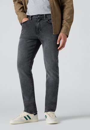 Homme portant un jean noir délavé, des baskets blanches avec des bandes vertes, une chemise grise et une veste marron avec une main dans la poche.