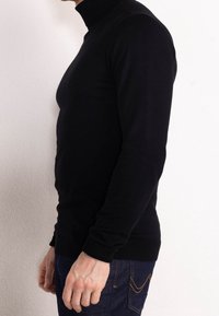 Schwarzer Rollkragenpullover aus dehnbarem Stoff, taillierte Passform mit langen Ärmeln und gerippten Bündchen, kombiniert mit dunklen Jeans.