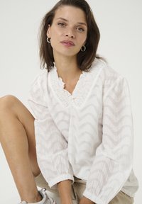 Blouse blanche avec un motif zigzag texturé, un col froncé et des manches bouffantes. Portée avec un bas rayé de couleur claire et des baskets.