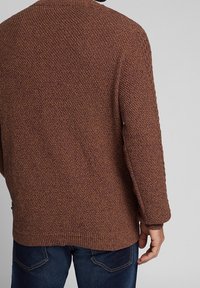 Brauner gestrickter Pullover mit einem strukturierten Muster, gerippten Bündchen und Saum. Der Rücken hat eine lockere Passform und wird mit blauen Jeans kombiniert.