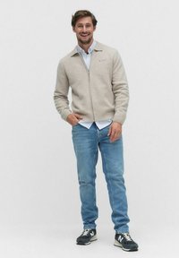 Beige zip-sweater med krage, bärs över en ljusblå skjorta med knappar, i kombination med ljusblå jeans och svarta sneakers.