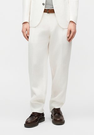 Hombre con pantalones blancos a medida, cinturón de cuero marrón y zapatos marrón oscuro de cuero con cordones, de pie contra un fondo blanco liso.