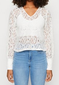 Blusa bianca in pizzo con maniche lunghe e scollatura a V, caratterizzata da motivi floreali, un design leggermente corto e una texture trasparente. Abbinata a jeans blu.