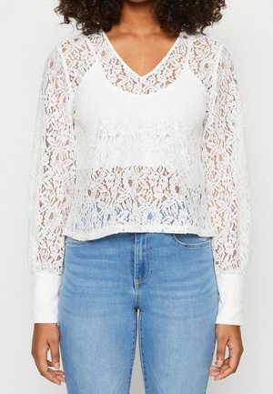 Weißes Spitzenbluse mit langen Ärmeln und V-Ausschnitt, mit Blumenmustern, einem leicht verkürzten Design und einer transparenten Textur. Kombiniert mit blauen Jeans.