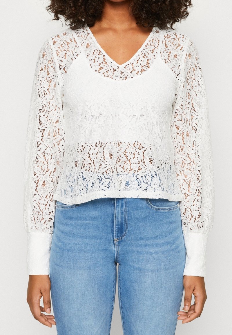 Blusa bianca in pizzo con maniche lunghe e scollatura a V, caratterizzata da motivi floreali, un design leggermente corto e una texture trasparente. Abbinata a jeans blu.