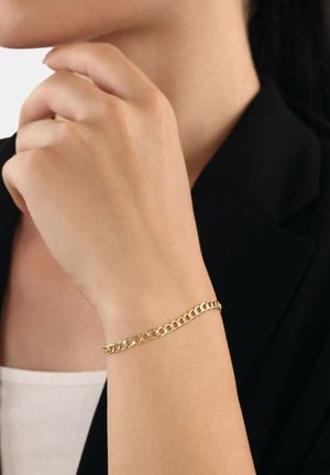 Gold chain bracelet on the wrist. The design features interlocking links with a smooth finish. The person wears a black blazer and white shirt.