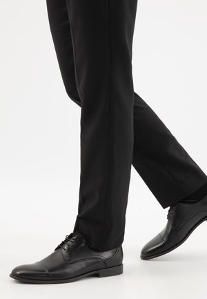 Schwarze Lederschuhe mit Kappenspitzen, getragen zu einer schwarzen Hose, gezeigt in einer Gehpose vor weißem Hintergrund.