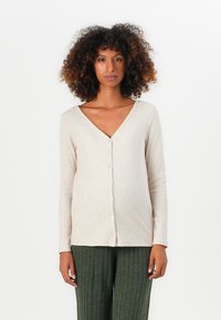 Kvinna med lockigt hår som bär en beige knappad V-ringad långärmad topp och gröna ribbade vida byxor, stående mot en enfärgad vit bakgrund.