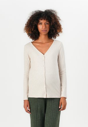 Kvinna med lockigt hår som bär en beige knappad V-ringad långärmad topp och gröna ribbade vida byxor, stående mot en enfärgad vit bakgrund.