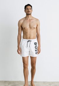 Bijele plivačke šortse s crnim logotipom "BOSS" na lijevoj strani, s pojasom na vezanje i laganom tkaninom.