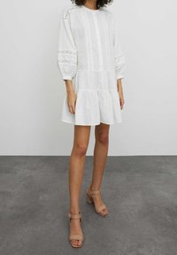 Robe en coton blanc avec manches longues bouffantes, fermeture par boutons et détails en dentelle. Design à étages avec une silhouette fluide. Portée avec des sandales à talons beiges.