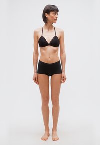 Γυναίκα με κοντά σκούρα μαλλιά, που φοράει μαύρο δαντελένιο σουτιέν και μαύρο σορτς, όρθια ξυπόλυτη μπροστά σε απλό λευκό φόντο, κοιτάζοντας δεξιά.