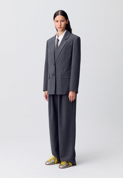 Traje gris a rayas con pantalones anchos y un blazer a medida. Camisa blanca y corbata negra a rayas. El calzado presenta detalles en plateado con acentos en amarillo.