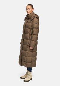 Brauner Puffer-Mantel mit Kapuze, langer Silhouette, gesteppter Optik, Reißverschluss vorne und Seitentaschen. Kombiniert mit beigen Schnürstiefeln und blauen Jeans.