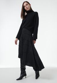 Zwarte coltrui met lange mouwen, gecombineerd met een gelaagde zwarte rok met verticale strepen, aangevuld met zwarte laarzen tot op de knie.