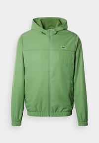 Zelena lahka jakna s kapuco, z zadrgo, elastičnimi manšetami in teksturiranim dizajnom. Na prsih je majhen logotip.