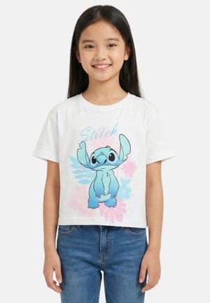 Giovane ragazza che sorride indossa una maglietta bianca con un personaggio Stitch blu e un motivo floreale rosa pastello e blu, abbinata a jeans blu.