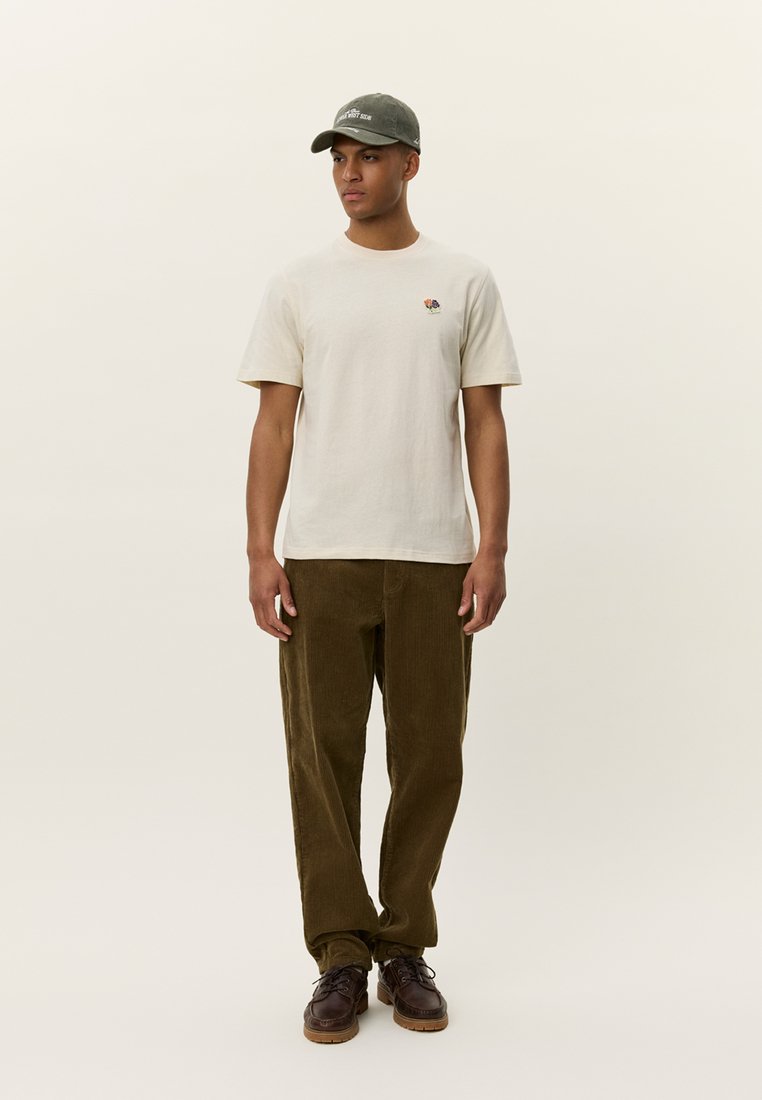 Beige cotton t-shirt with small colorful embroidered detail, paired with dark olive corduroy trousers and brown leather shoes, styled with a green cap.