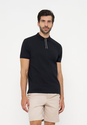 Mand med mørkt hår og skæg iført en sort kortærmet polotrøje med en hvidstribet lynlås og beige shorts, der står mod en hvid baggrund.