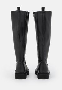 Botas negras hasta la rodilla hechas de un material sintético suave, con cremalleras laterales, suela plana y un diseño cosido alrededor del tobillo.