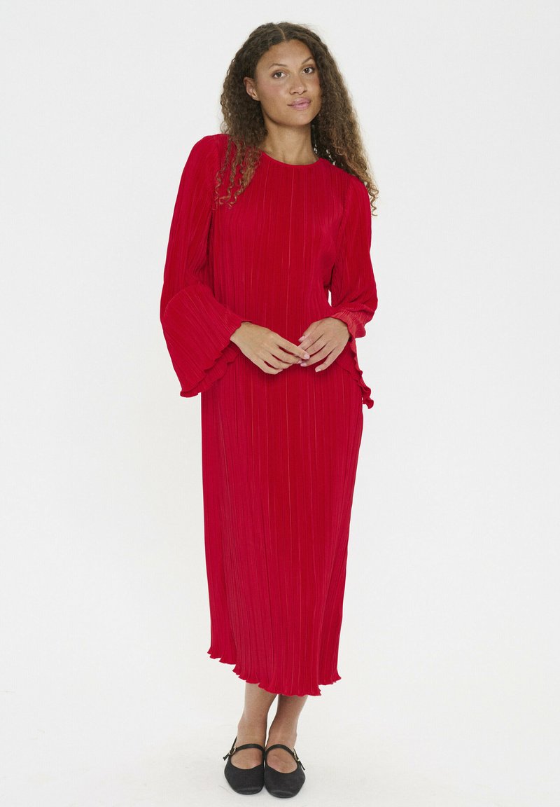 Rochie plisată roșie cu mâneci lungi și evazate și accente volănașe, reaching the mid-calf, realizată dintr-un material ușor. Pantofii negri cu talpă joasă completează aspectul.