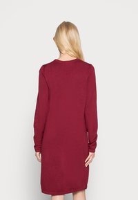 Blonde vrouw staat met haar rug naar de camera gekeerd, gekleed in een lang mouwend, knielang bordeauxrode trui jurk tegen een witte achtergrond.