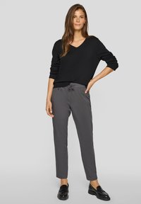 Schwarzer V-Ausschnitt-Pullover, weiche Strickstruktur; graue, zulaufende Hose mit Kordelzug in der Taille und seitlichen Taschen; getragen mit schwarzen Loafers.