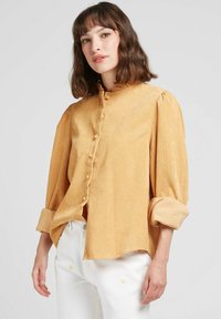 Chemise boutonnière jaune moutarde avec col froncé, texture douce et manches bouffantes. Portée avec un pantalon blanc aux accents floraux.