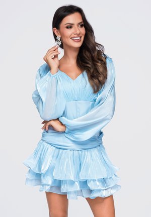 Femme souriante touchant de grandes boucles d’oreilles florales, portant une robe bleu clair à manches longues avec jupe à volants superposés.