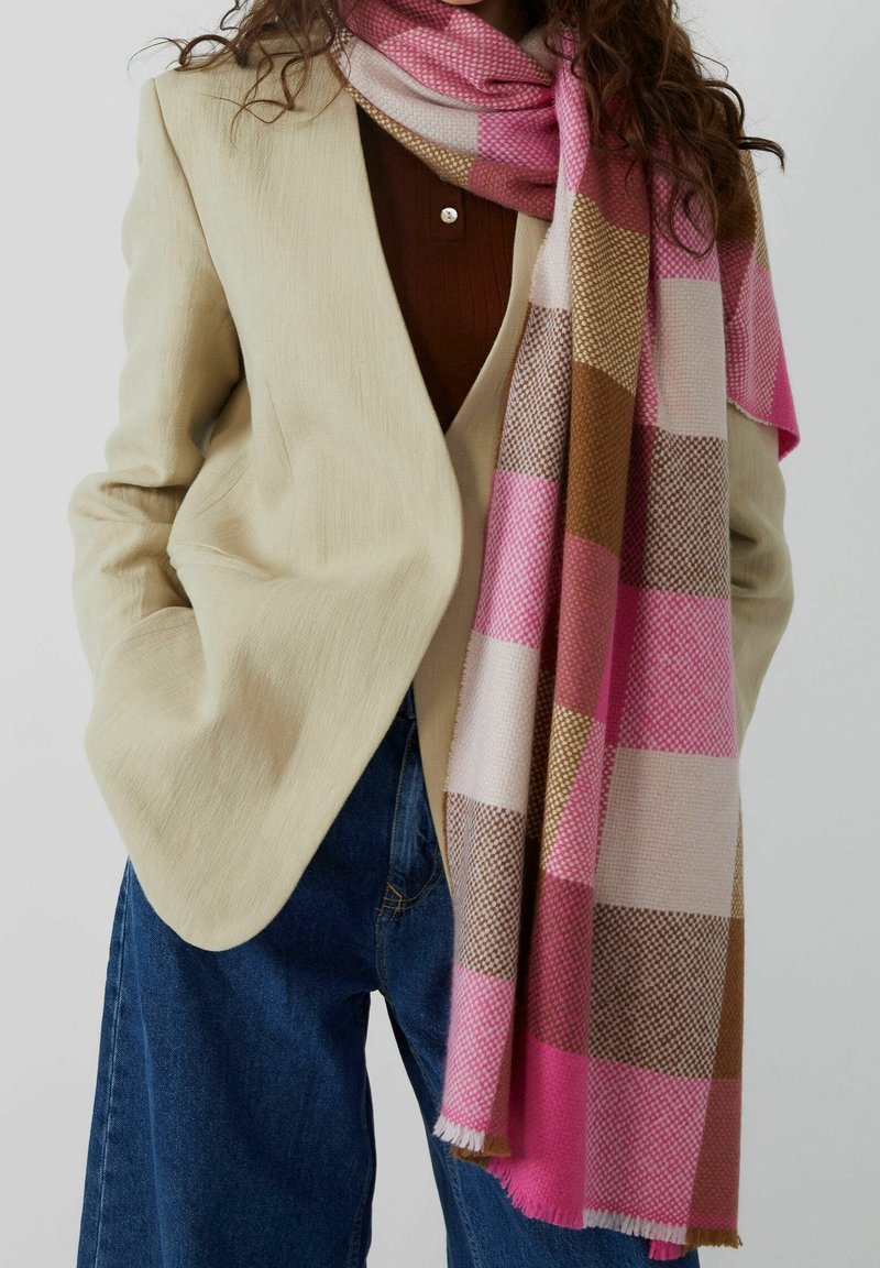 Femme portant un blazer beige, un top marron, un jean bleu et une longue écharpe à carreaux rose, marron et blanc drapée autour du cou.