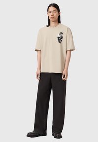 Camiseta de algodón beige con mangas cortas y un diseño gráfico de calavera en el pecho izquierdo, combinada con pantalones negros de corte holgado y zapatos negros.