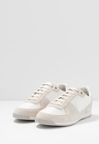 Bílé a béžové sportovní boty s kombinací semišového a síťovaného materiálu, kulatou špičkou, šněrováním vpředu a jemným logem značky.